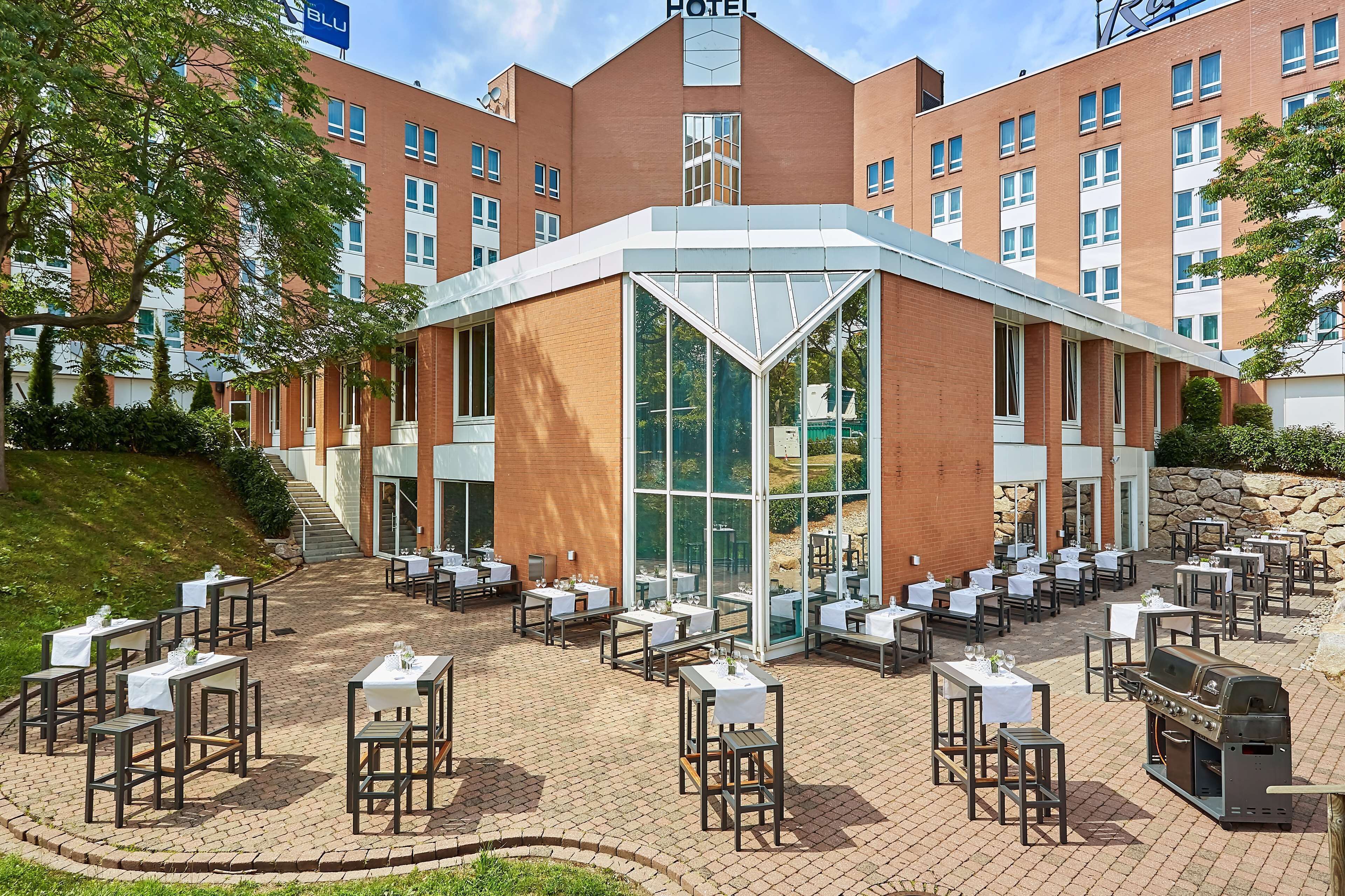 A hotel with a central glass atrium, outdoor dining area with tables and chairs, and a brick building facade.