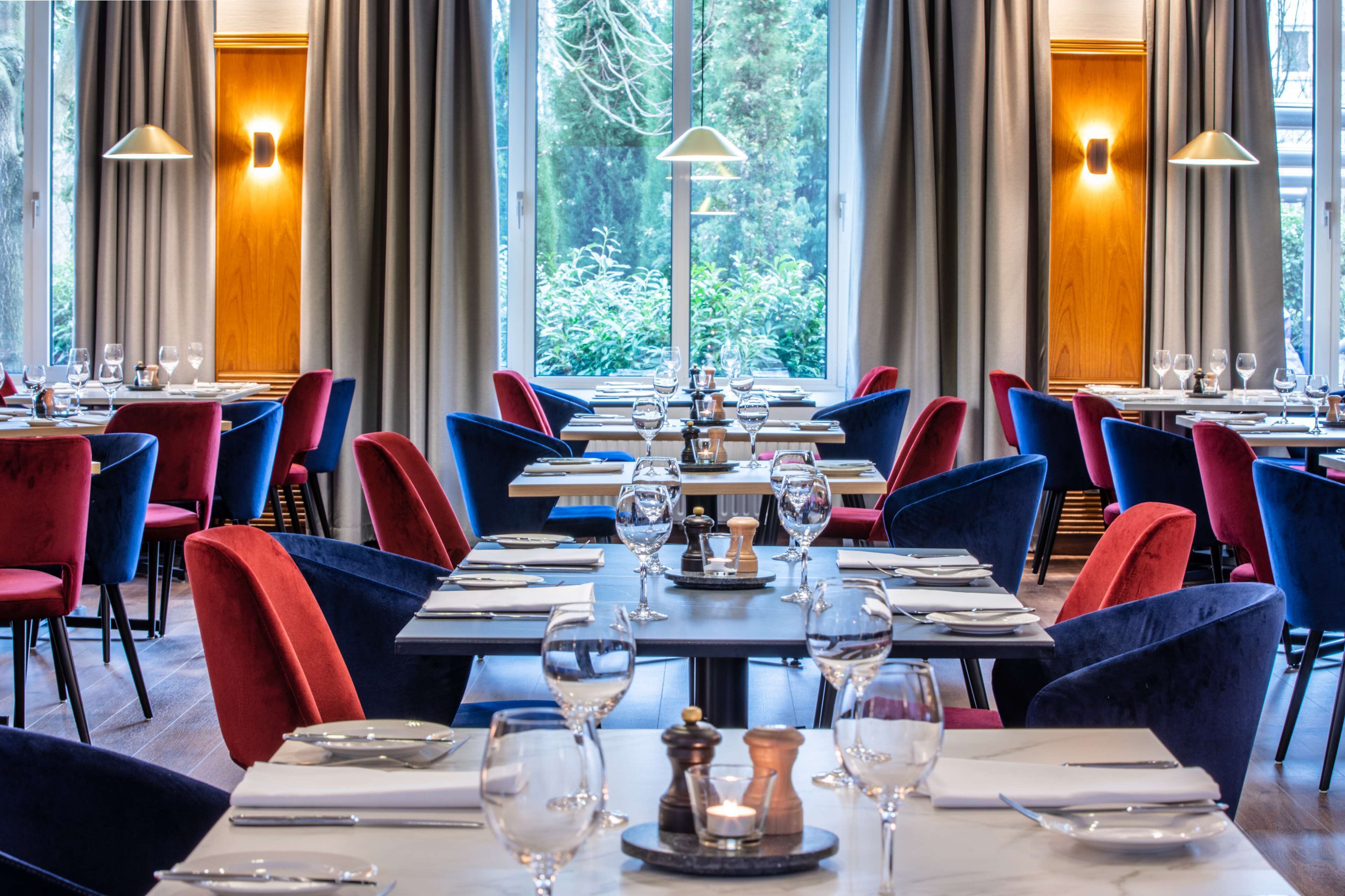 Un restaurante con mesas dispuestas para comer, sillas rojas y azules, y grandes ventanales con cortinas.