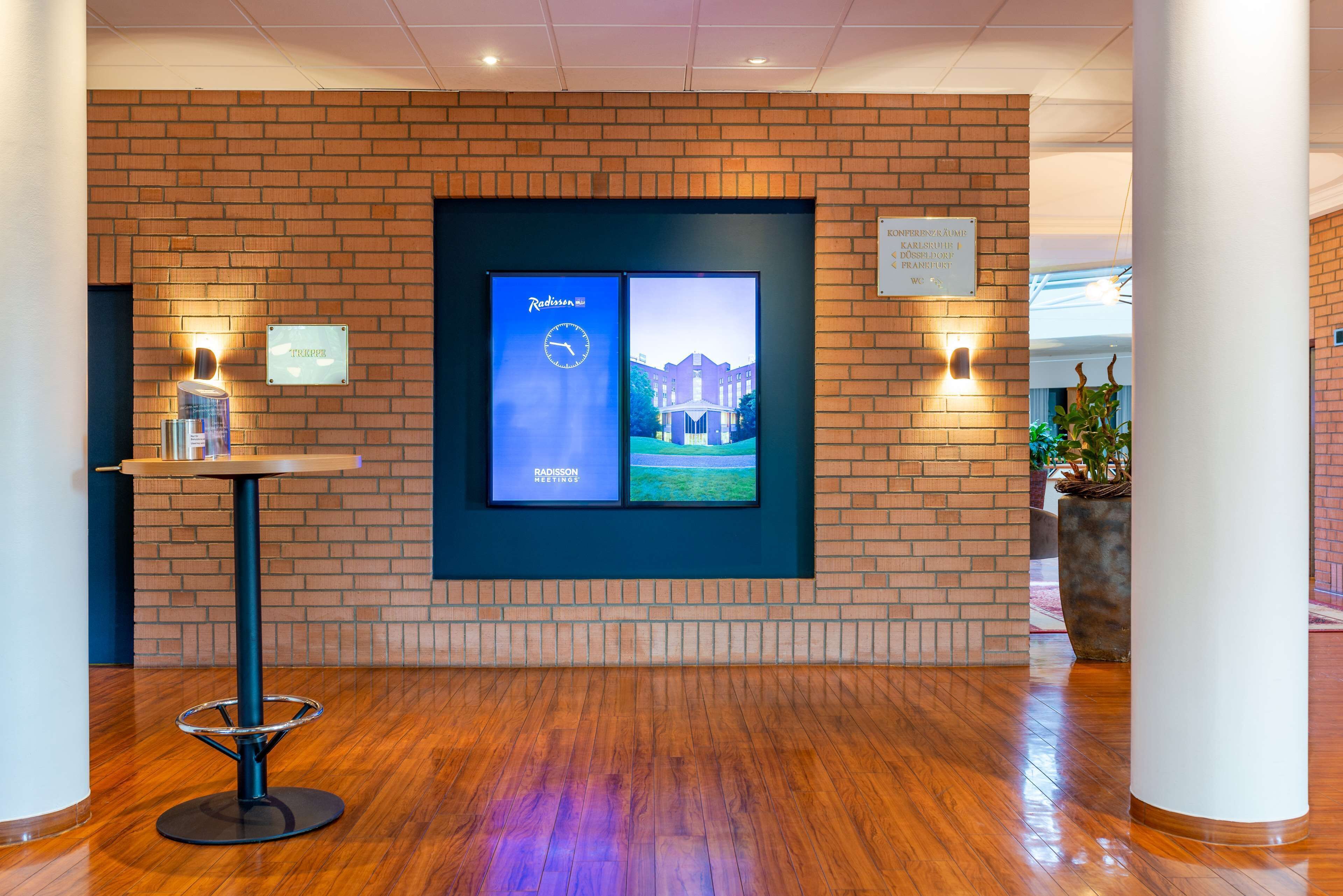 Een hal met een bakstenen muur, een digitaal scherm met Radisson Meetings branding, een kleine tafel met een fles en een potplant.