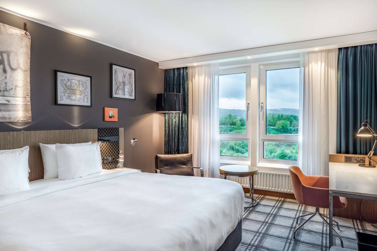 Habitación de hotel con una cama grande, un escritorio, una silla y vistas a zonas verdes a través de la ventana.