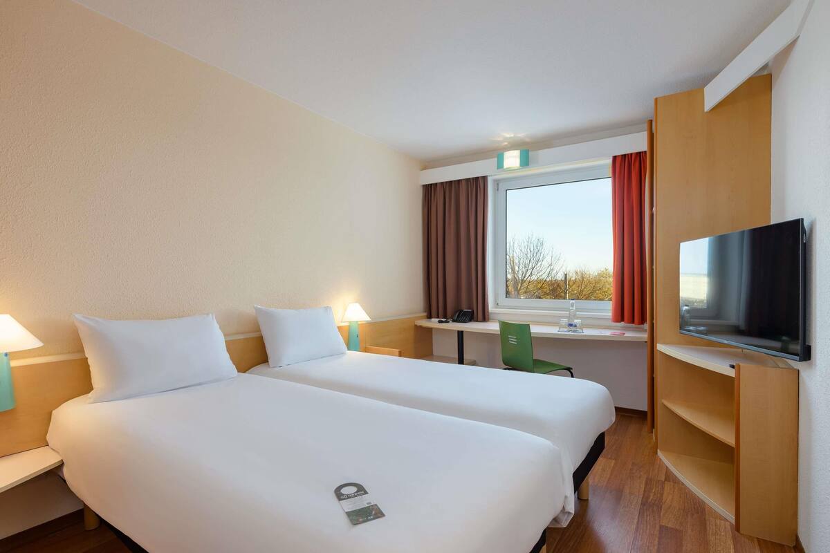 Habitación de hotel con dos camas, un escritorio, una silla, un televisor y una ventana con cortinas.