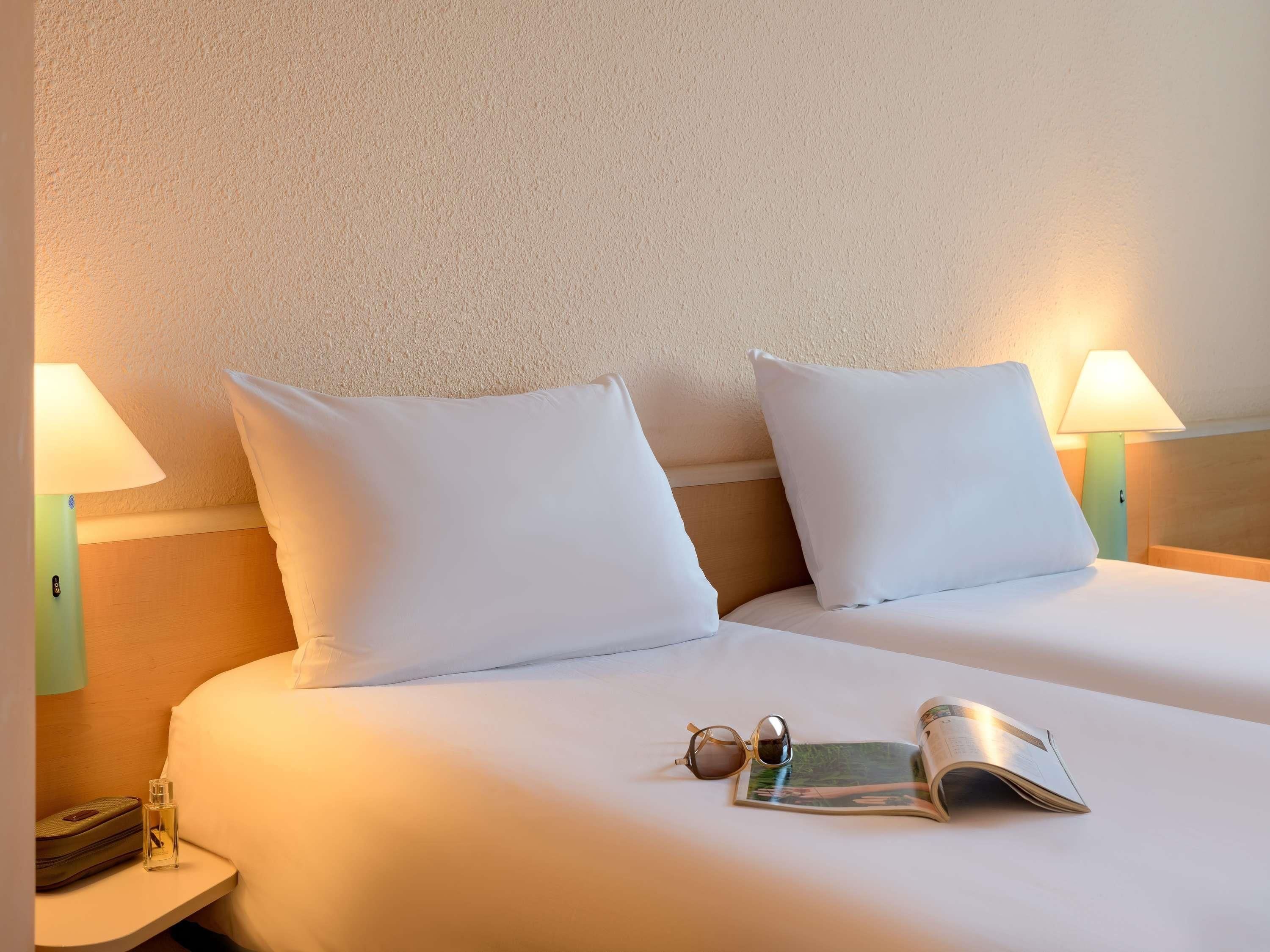 Et hotelværelse med to senge, begge med hvide sengetøj og puder, et natbord med en lampe, et par solbriller og et magasin.