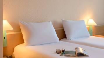 Ein Hotelzimmer mit zwei Betten, jeweils mit weißer Bettwäsche und Kissen, einem Nachttisch mit Lampe, einer Sonnenbrille und einer Zeitschrift.