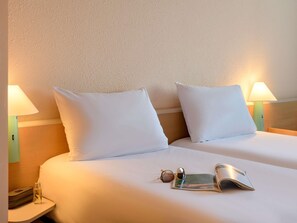 Une chambre d’hôtel avec deux lits, chacun agrémenté de draps et d’oreillers blancs, une table de chevet avec une lampe, une paire de lunettes de soleil et un magazine.