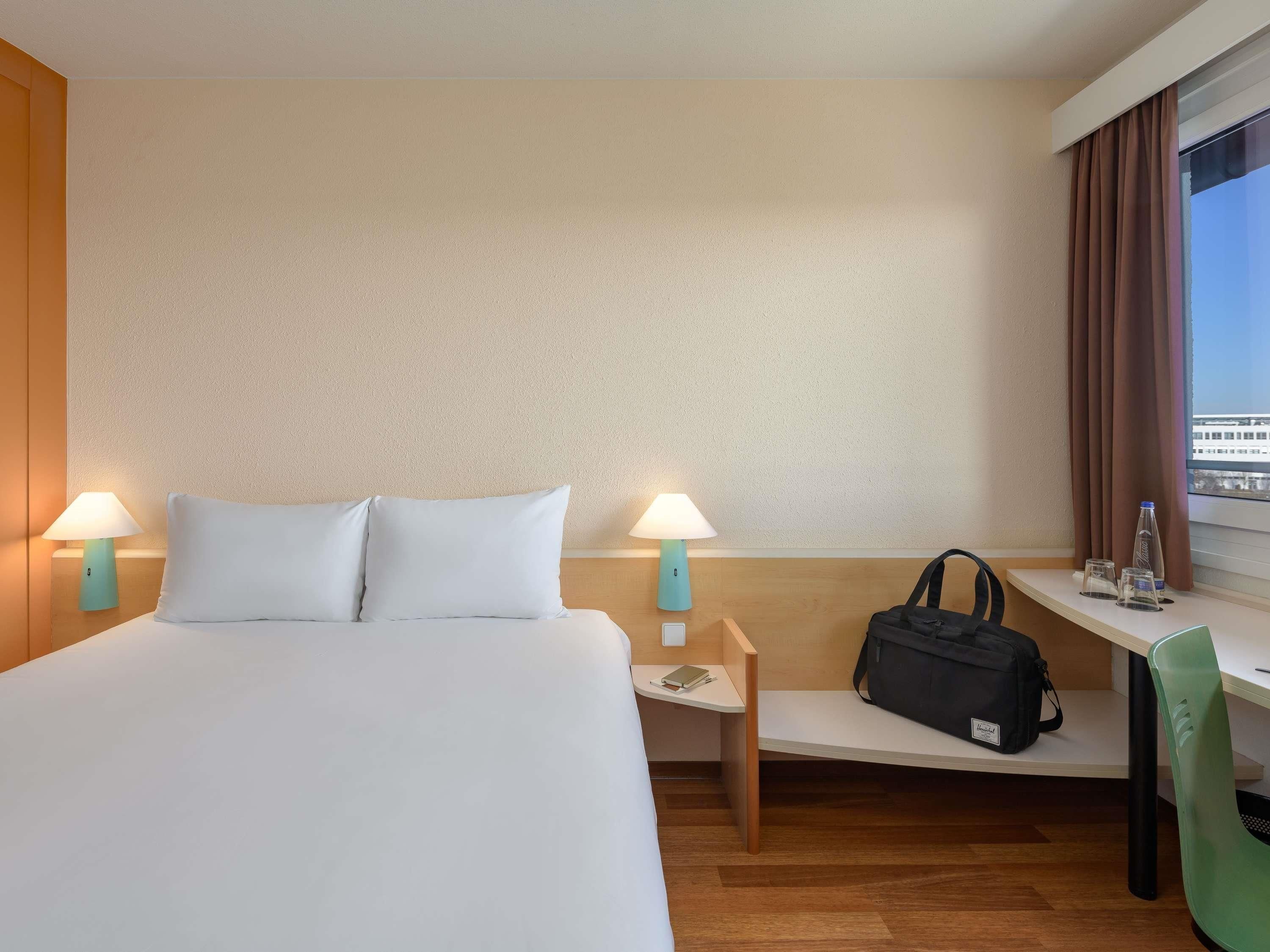 A hotel room with a bed, two bedside lamps, a desk with a chair, a window with curtains, and a bag on the desk.