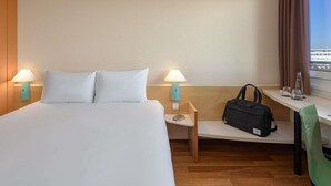 Habitación de hotel con una cama, dos lámparas de noche, un escritorio con una silla, una ventana con cortinas y un bolso sobre el escritorio.