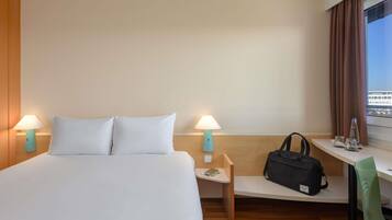 A hotel room with a bed, two bedside lamps, a desk with a chair, a window with curtains, and a bag on the desk.
