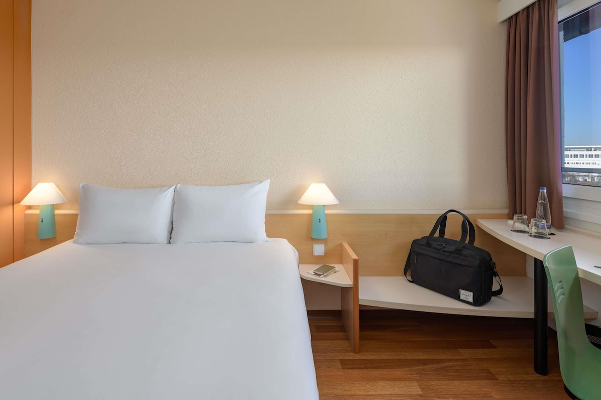 Habitación de hotel con una cama, dos lámparas de noche, un escritorio con una silla, una ventana con cortinas y un bolso sobre el escritorio.