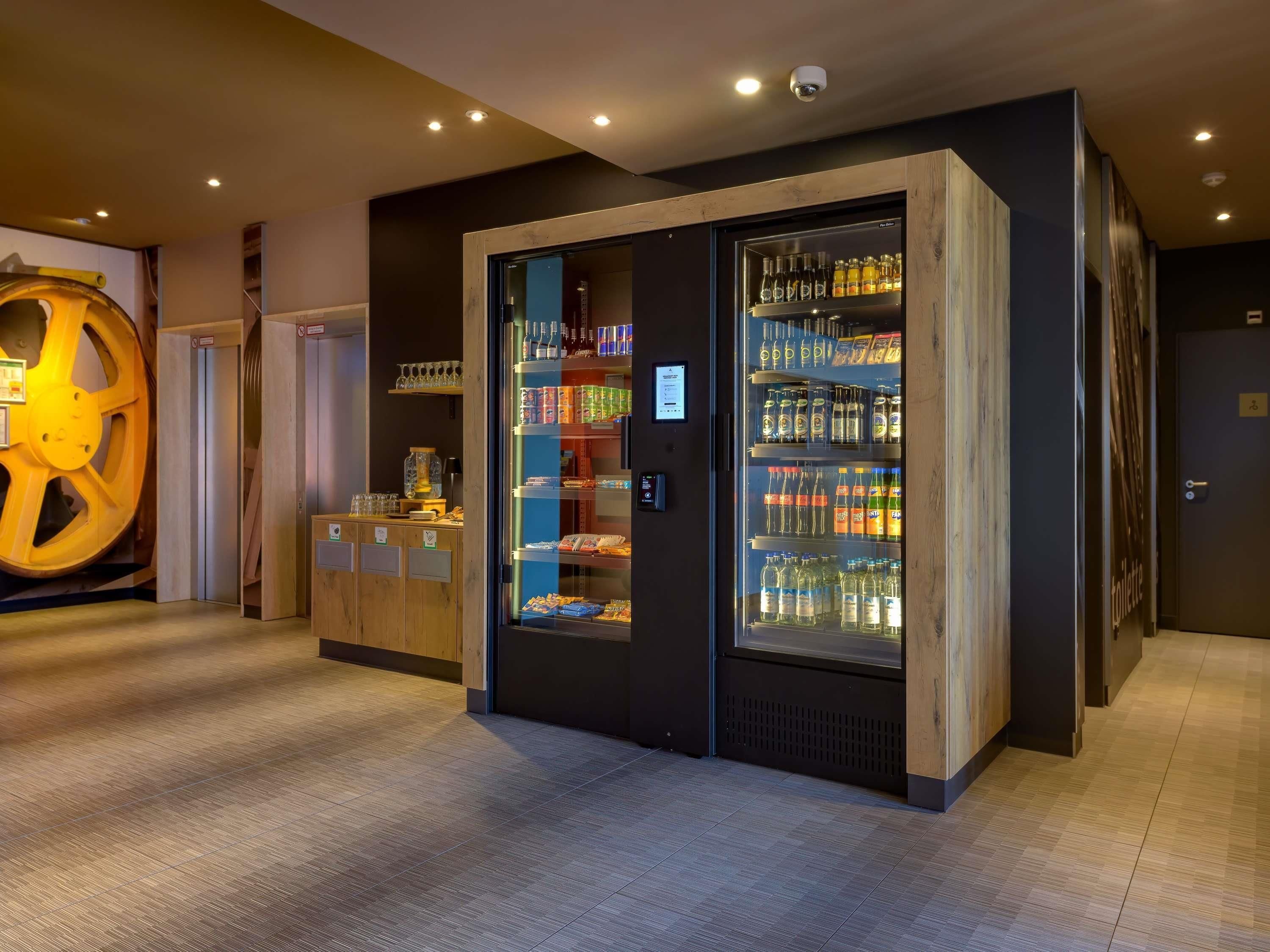 A modern hotel lobby with a large, illuminated sculpture, a self-service beverage station, and a refrigerated display case with various drinks.