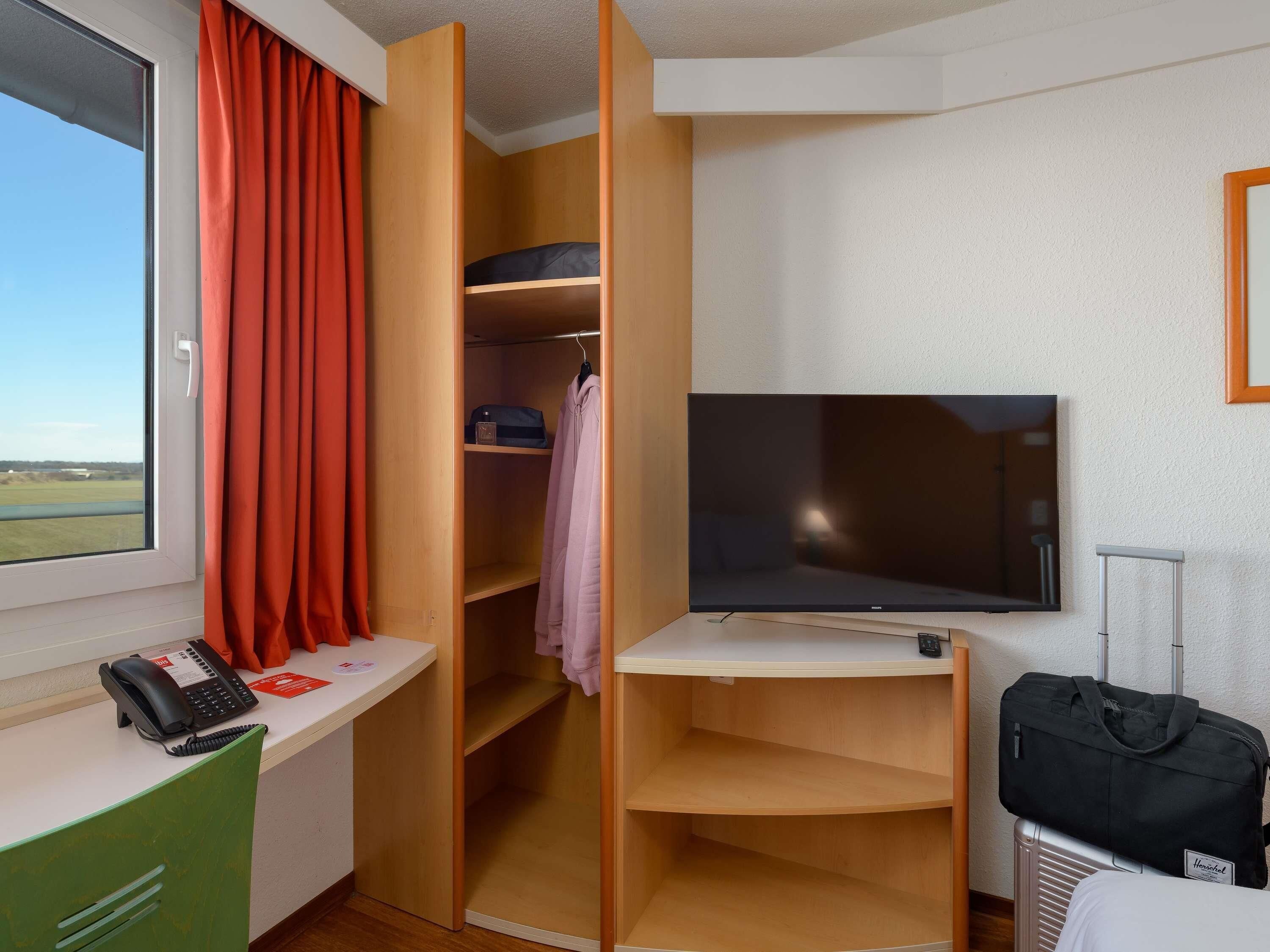 Ein Hotelzimmer mit einem Bett, einem Schreibtisch mit Telefon, einem Fernseher und einem Kleiderschrank mit Regalen.