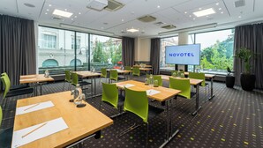 A hotel room with a large window, a TV screen displaying "NOVOTEL", and a conference room with tables and chairs. - Novotel Vilnius Centre (Vilnius)