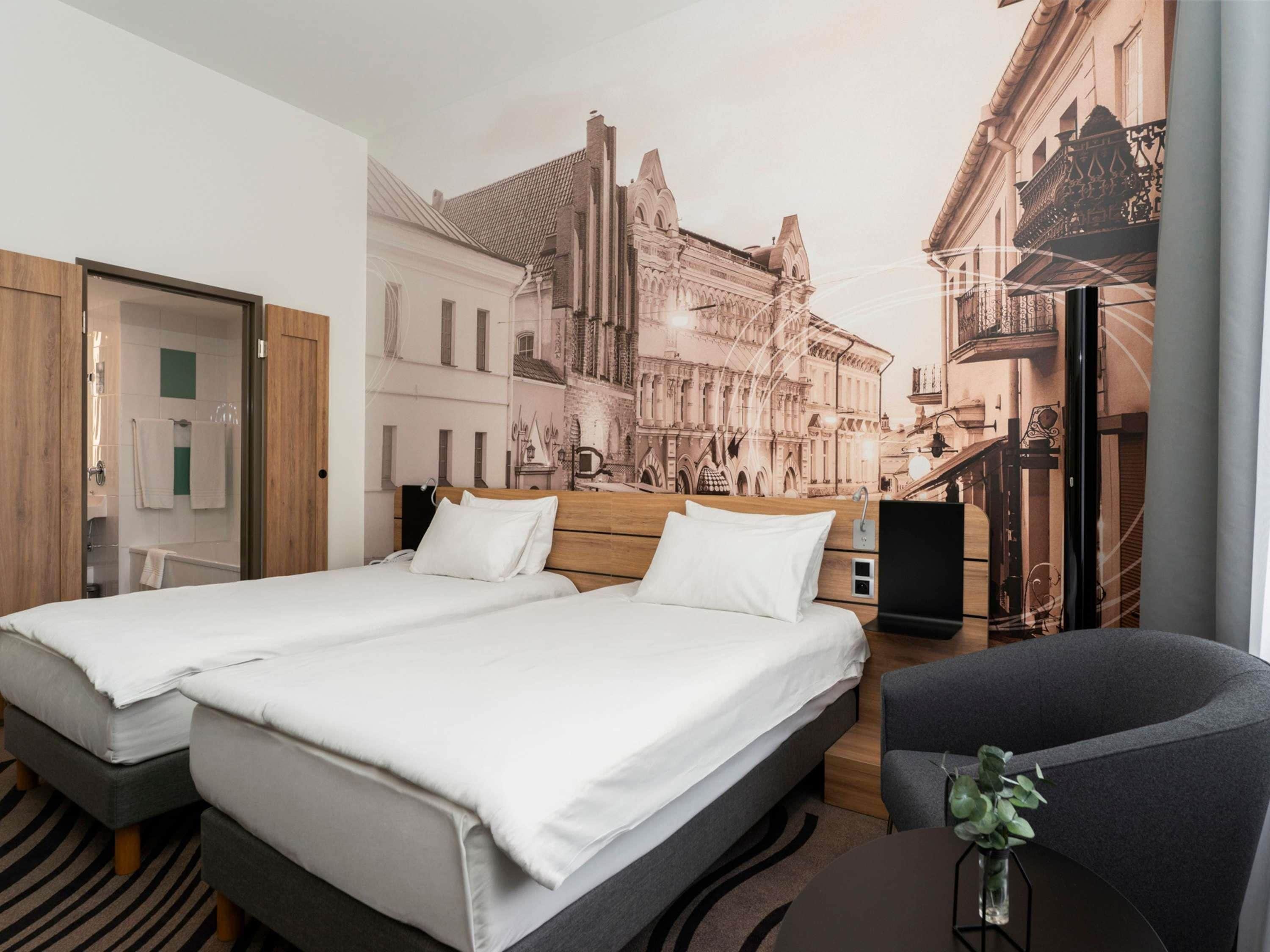 Habitación de hotel con cama doble, una silla, una mesita, televisión y un mural con un paisaje urbano.