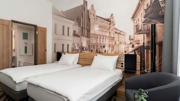 Habitación de hotel con cama doble, una silla, una mesita, televisión y un mural con un paisaje urbano.