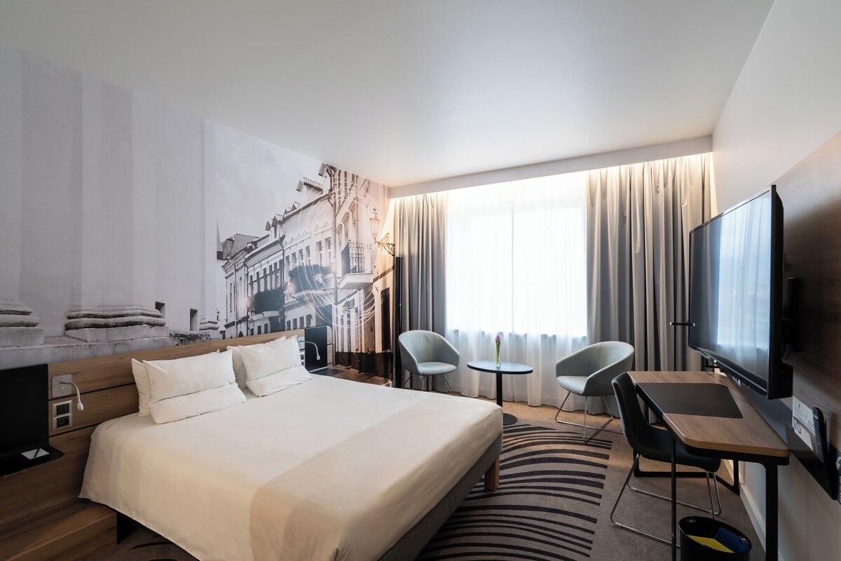 A hotel room with a large bed, a desk, two chairs, a flat-screen TV, and a cityscape mural on the wall.