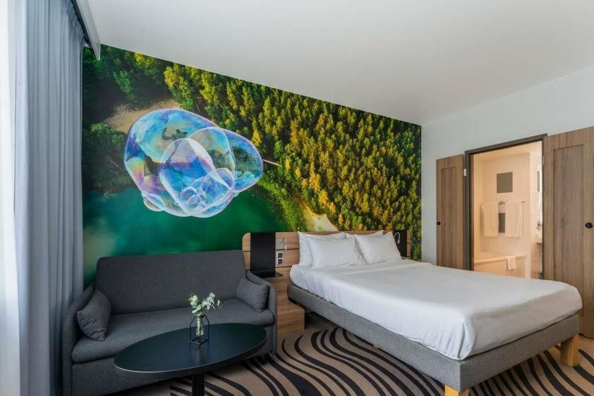 A modern hotel room with a bed, sofa, and a wall mural.