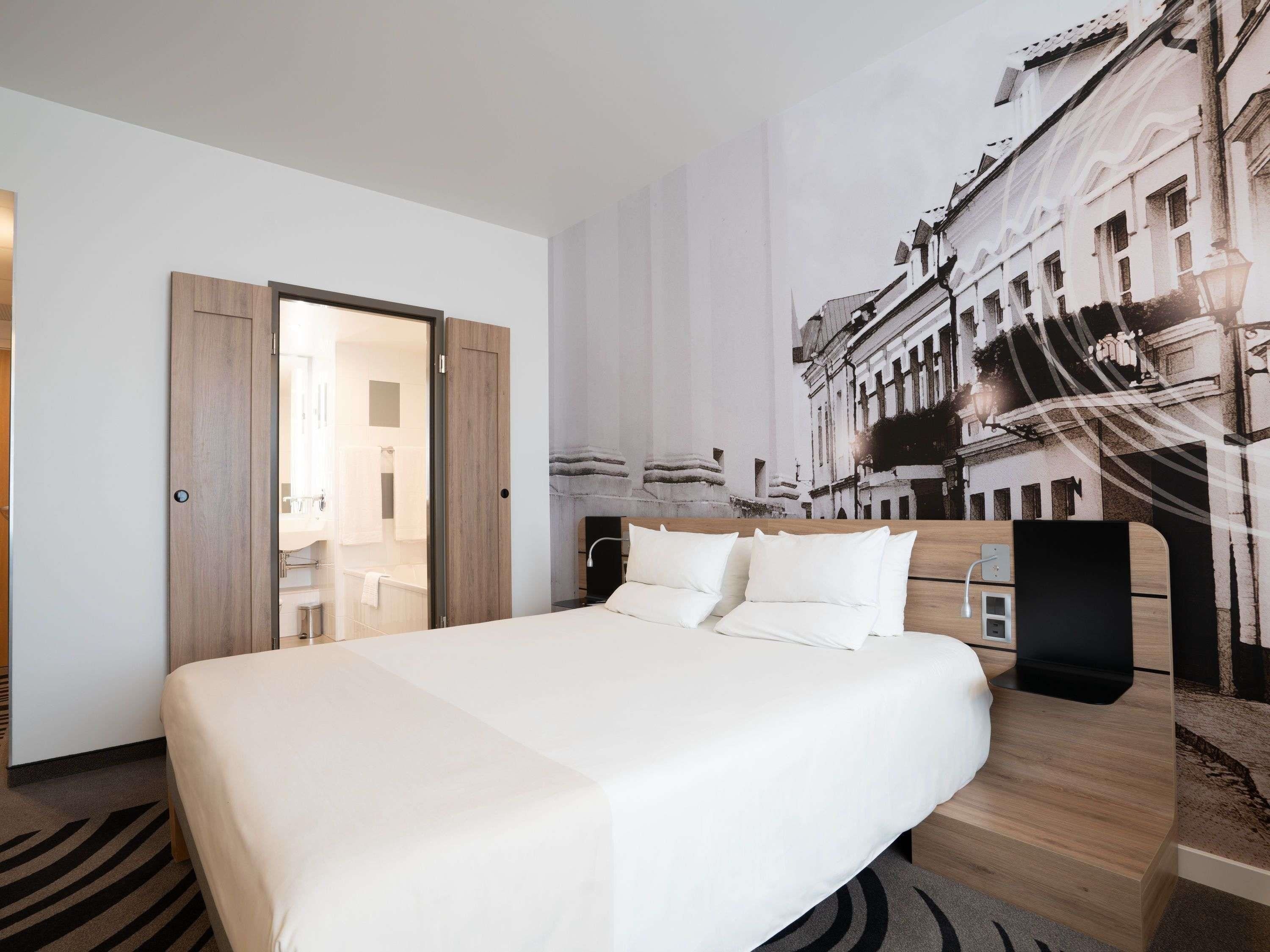 A hotel room with a large bed, a wooden headboard, a wall-mounted TV, and a cityscape mural.