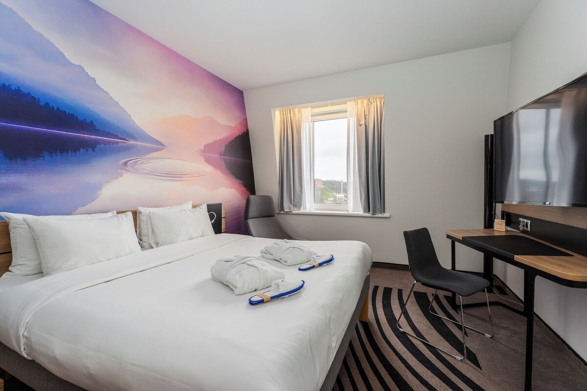 A hotel room with a bed, a desk, a chair, a TV, and a window with curtains.