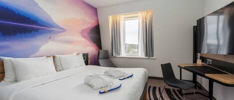 A hotel room with a bed, a desk, a chair, a TV, and a window with curtains.