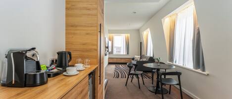 A modern hotel room with a kitchenette, a dining area, and large windows with curtains.