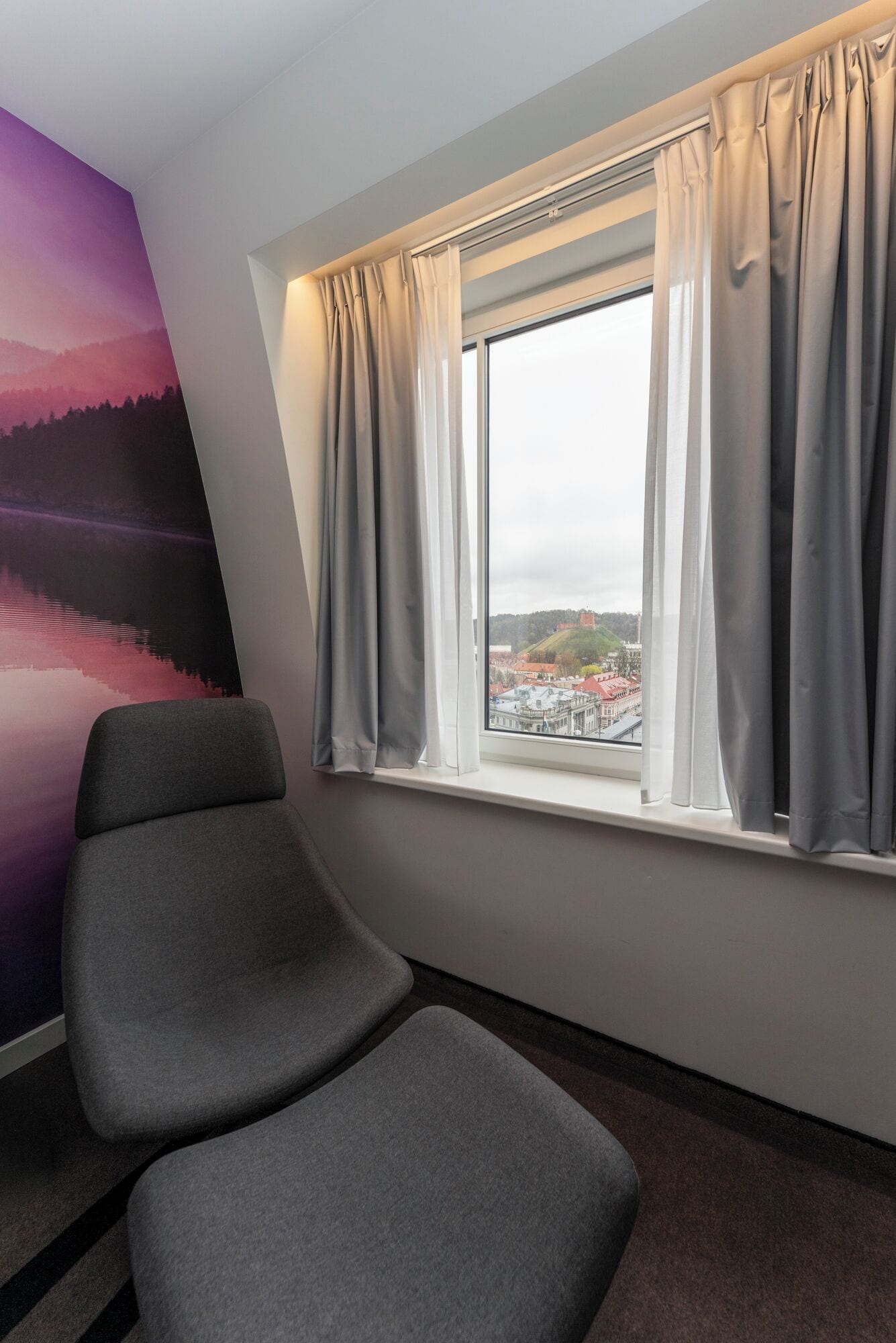 Una habitación con un ventanal grande, una silla gris y un mural en la pared.