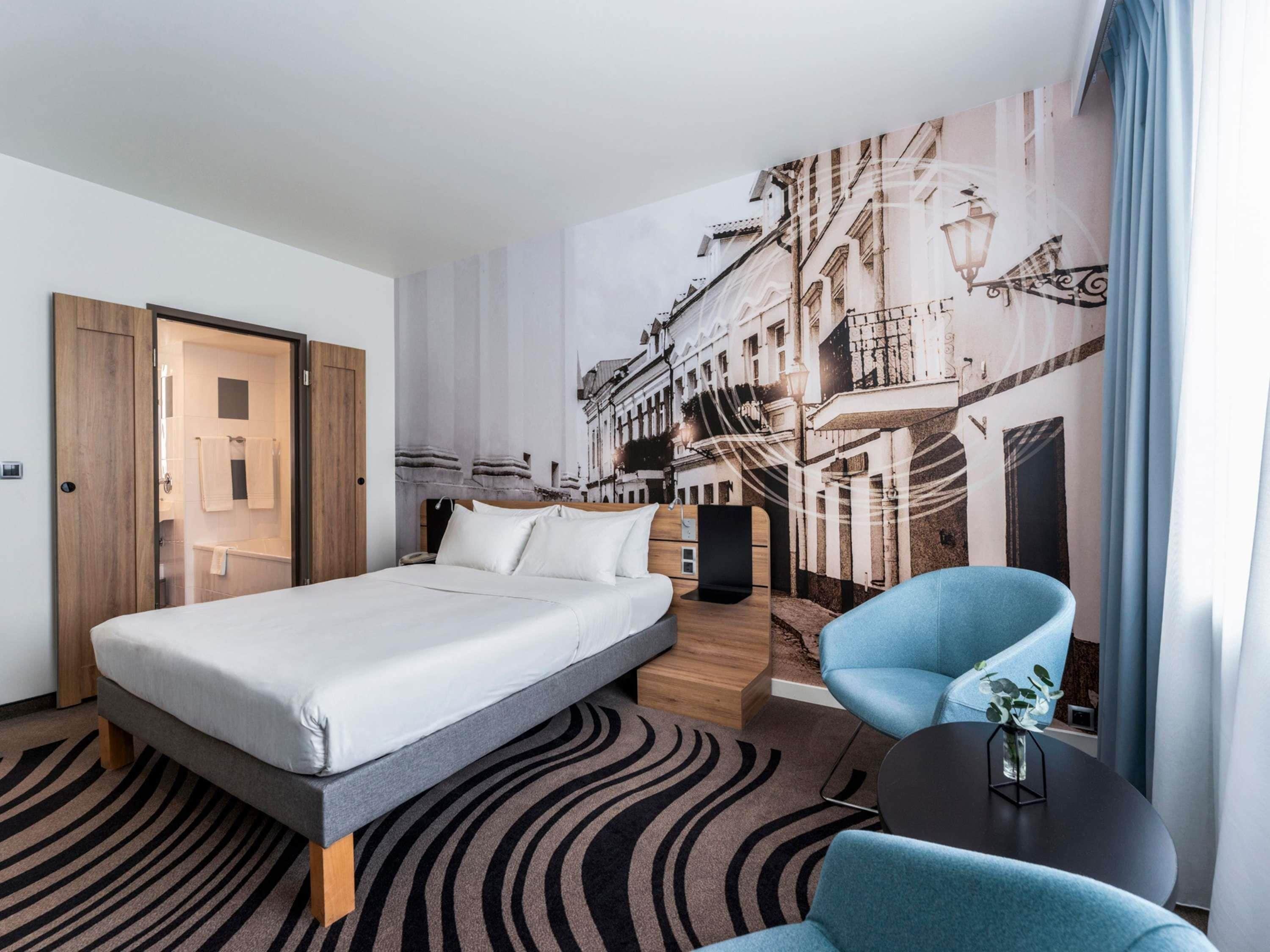 A hotel room with a bed, a chair, a small table, and a wall mural.