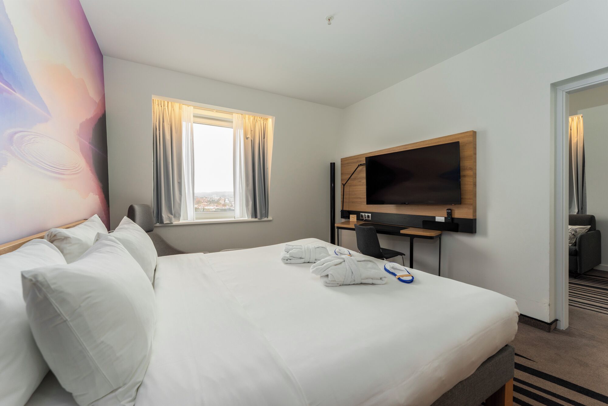 Una habitación de hotel con una cama, televisión, escritorio y ventana con cortinas.
