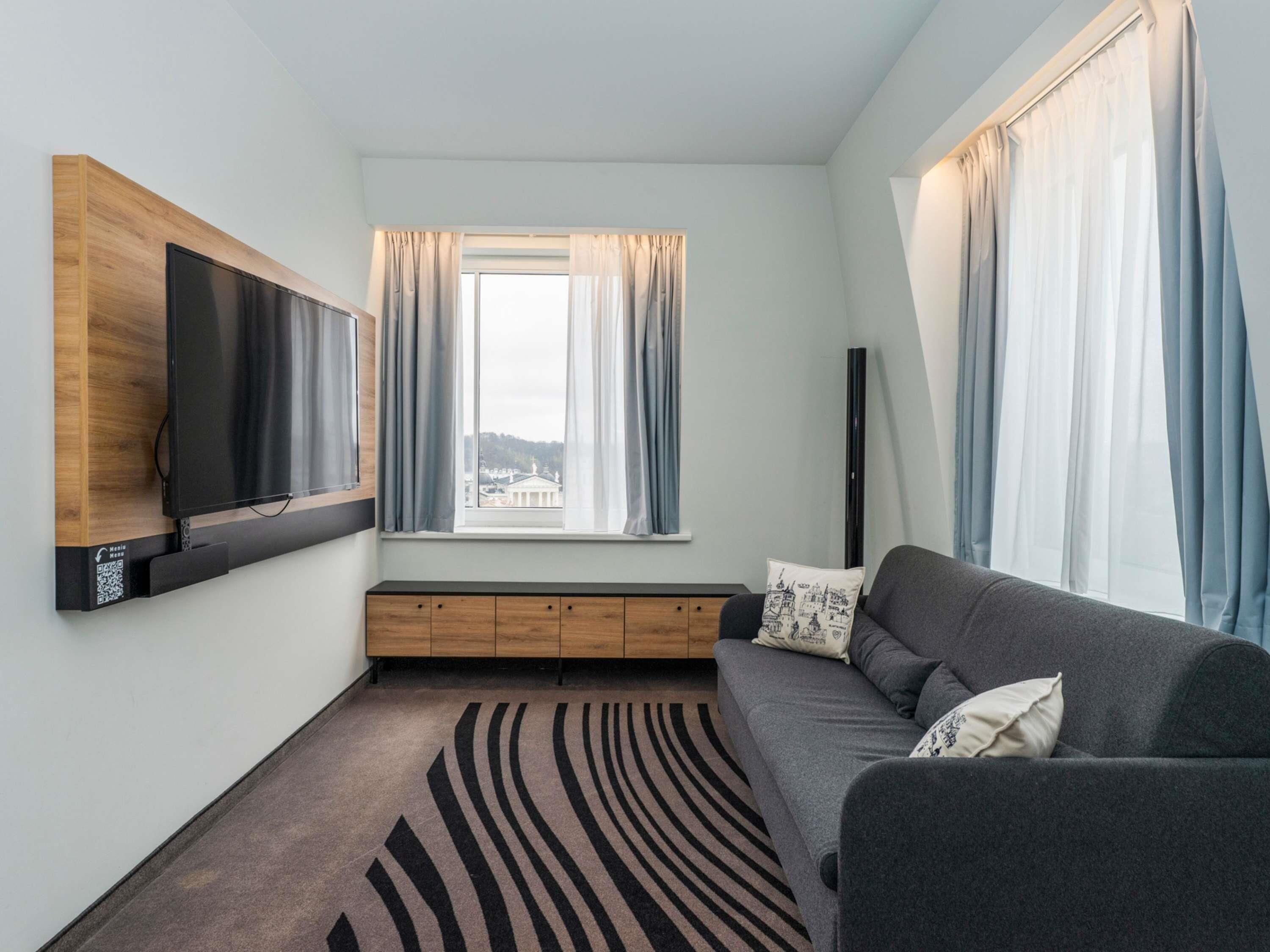 Un salon moderne avec un téléviseur à écran plat fixé sur un panneau en bois, un canapé gris agrémenté de coussins à motifs, une fenêtre avec des rideaux légers et un tapis à motifs.