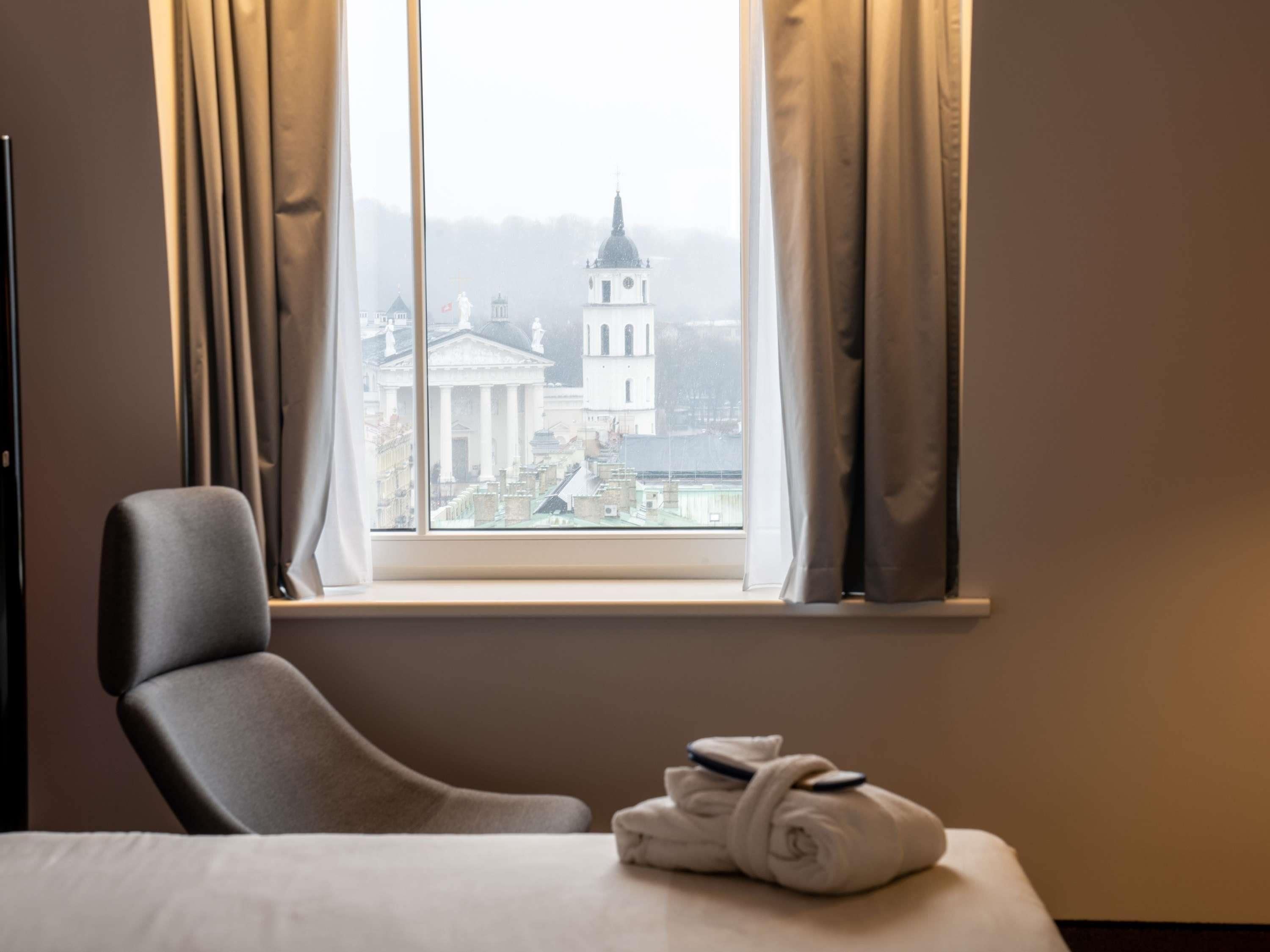 A hotel room with a bed, a chair, a window with curtains, and a view of a building with a dome.