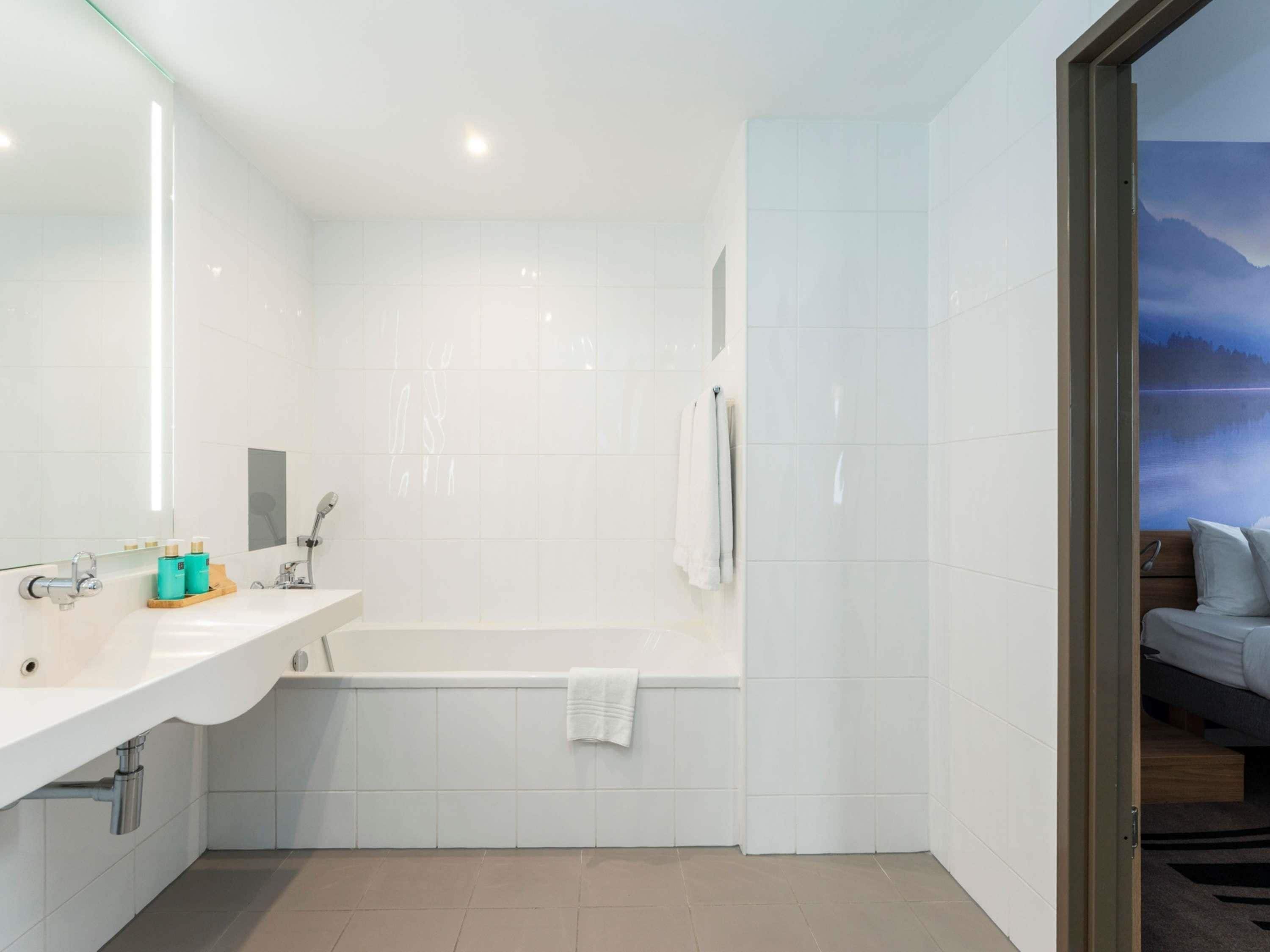 Une salle de bain moderne avec une grande baignoire, un meuble-lavabo avec un miroir et une vue sur un lac, visible à travers une porte ouverte.