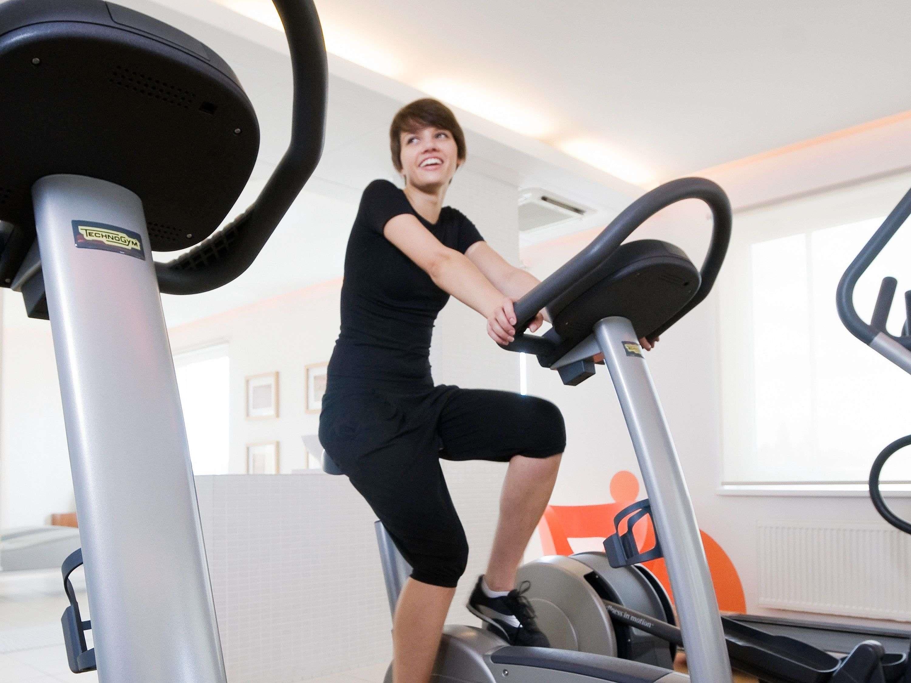 Una persona usando una bicicleta estática en un gimnasio con una pared blanca y una ventana al fondo.