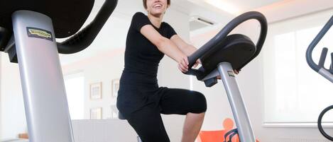 Una persona usando una bicicleta estática en un gimnasio con una pared blanca y una ventana al fondo.