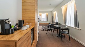 A modern hotel room with a wooden kitchenette, a dining area with a round table and chairs, and large windows with curtains. - Novotel Vilnius Centre (Vilnius)