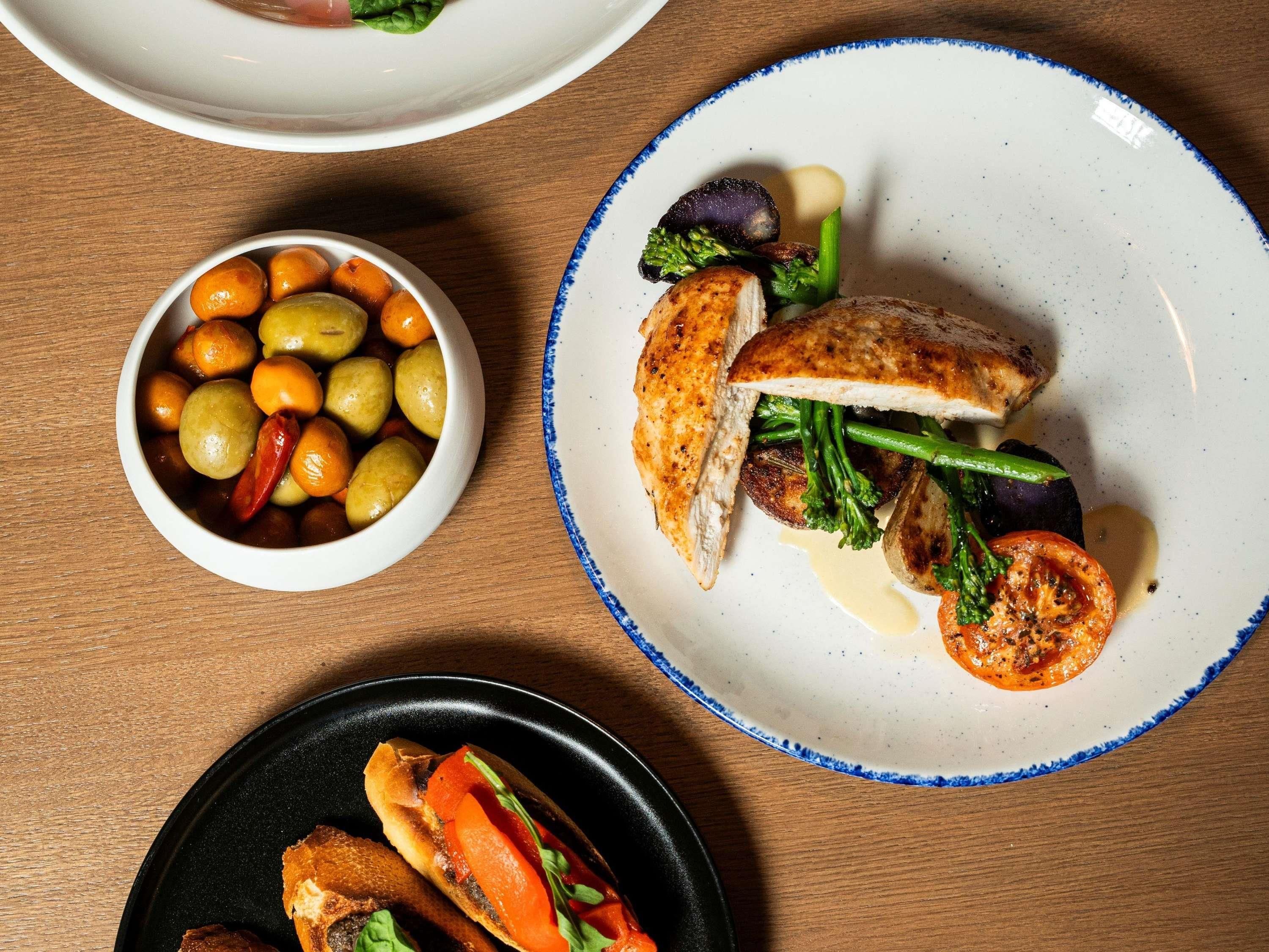 Una mesa con platos de comida, incluyendo una ensalada de hojas verdes y tomates, un bol de aceitunas y un plato con pechuga de pollo a la parrilla.