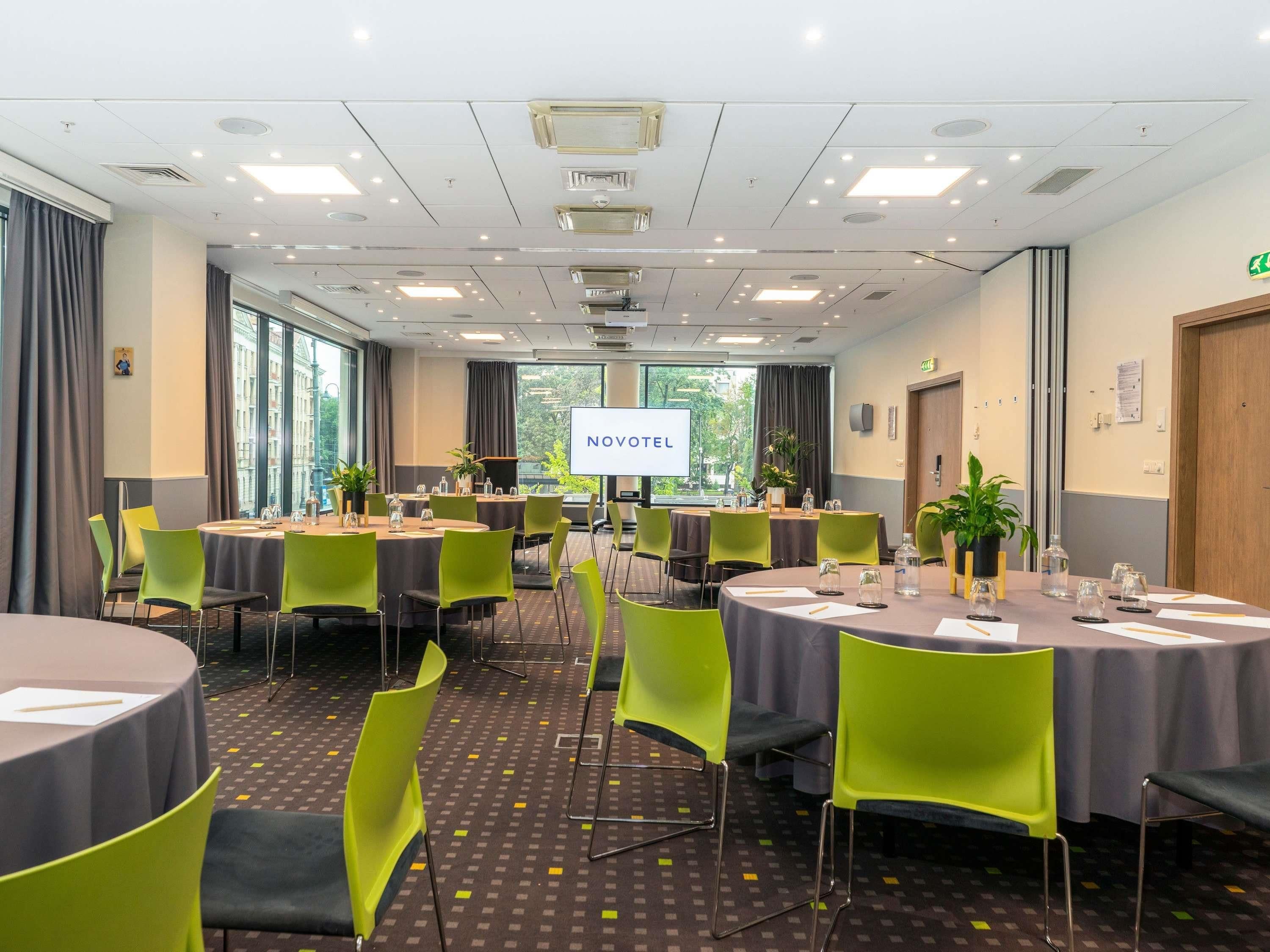 Ein Konferenzraum mit mehreren Tischen, Stühlen und einer Projektionsleinwand, die 'NOVOTEL' zeigt.