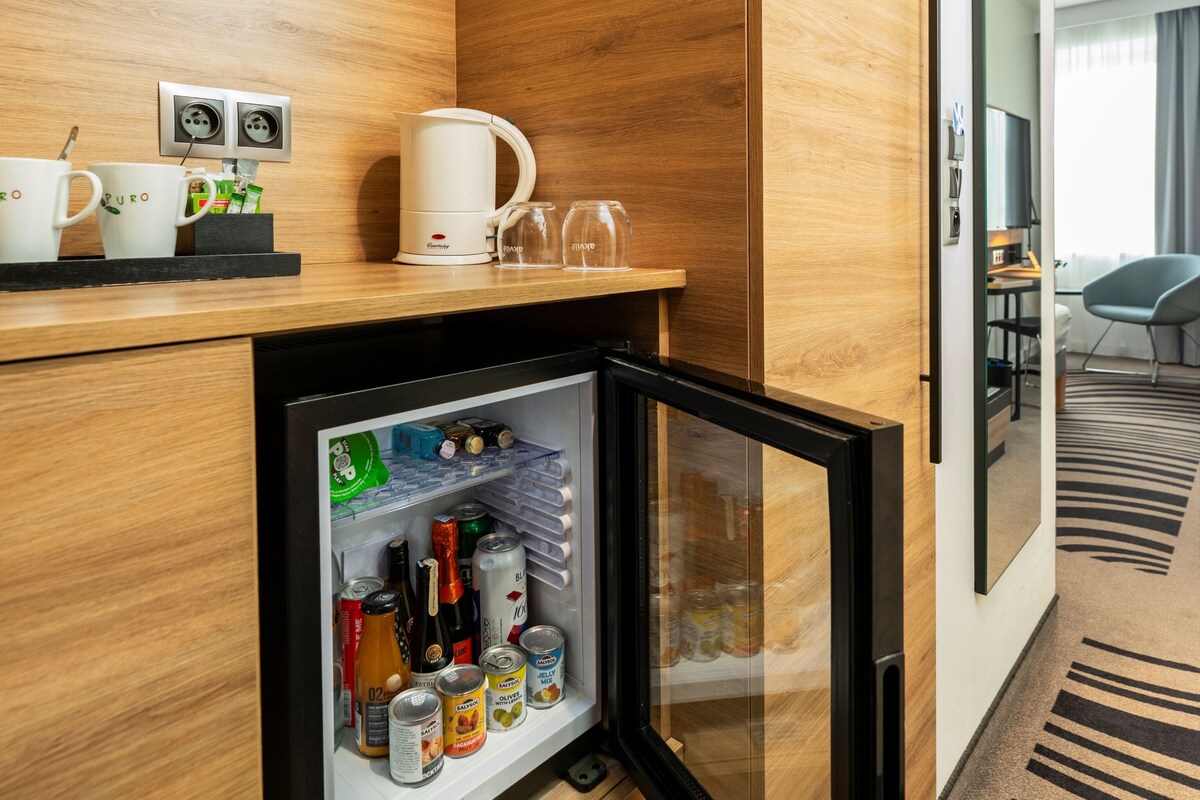 A hotel minibar with a refrigerator stocked with beverages and snacks, a coffee machine, and two cups on the counter.