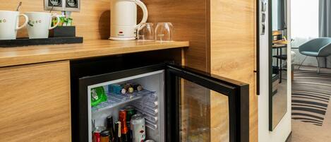 A hotel minibar with a refrigerator stocked with beverages and snacks, a coffee machine, and two cups on the counter.