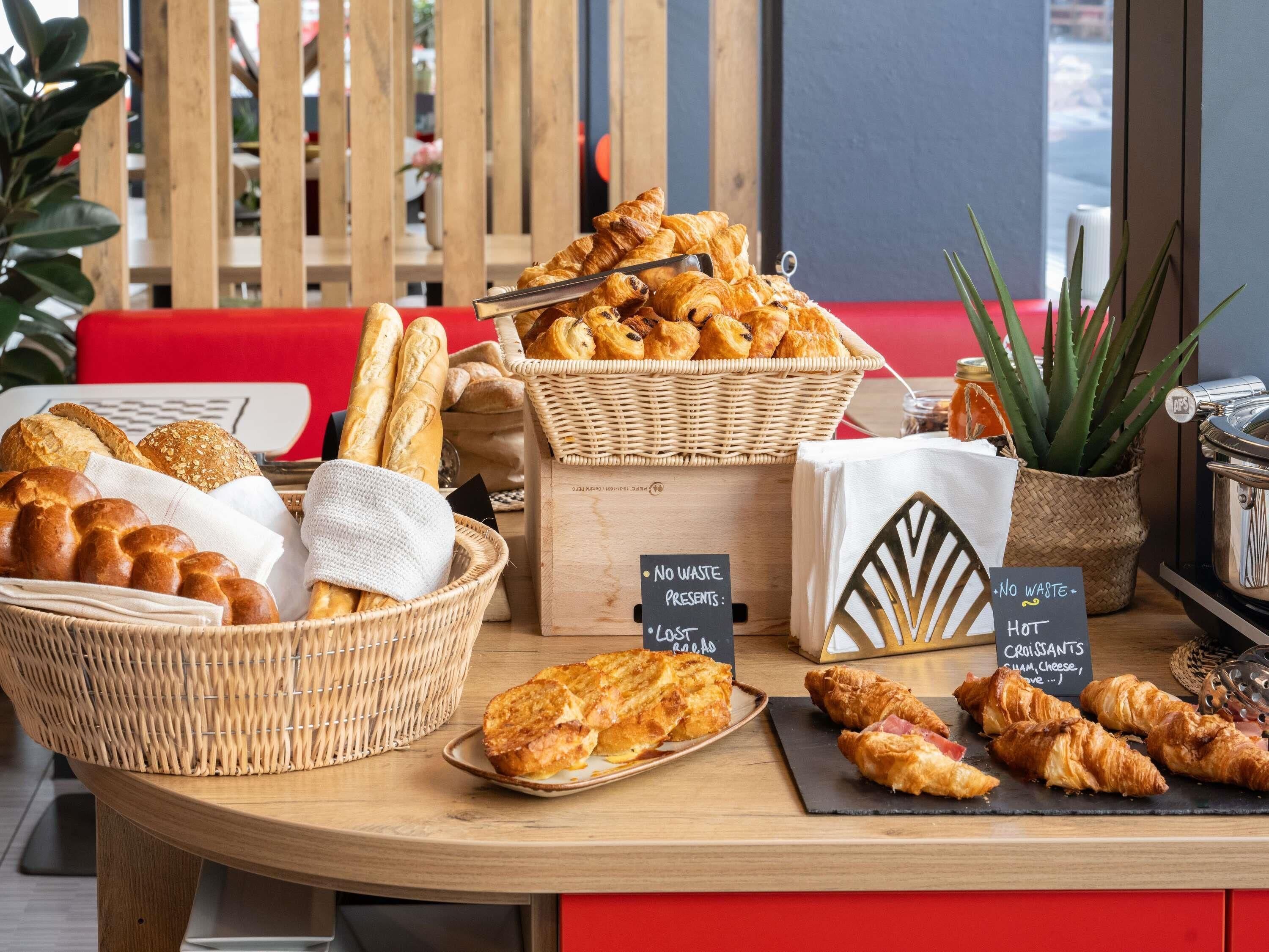A variety of baked goods displayed on a wooden table, including bread, croissants, and pastries, with signs indicating no waste and hot options.