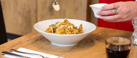 A bowl of pasta with a spoonful being added, a glass of dark liquid, and a napkin with utensils on a wooden table.