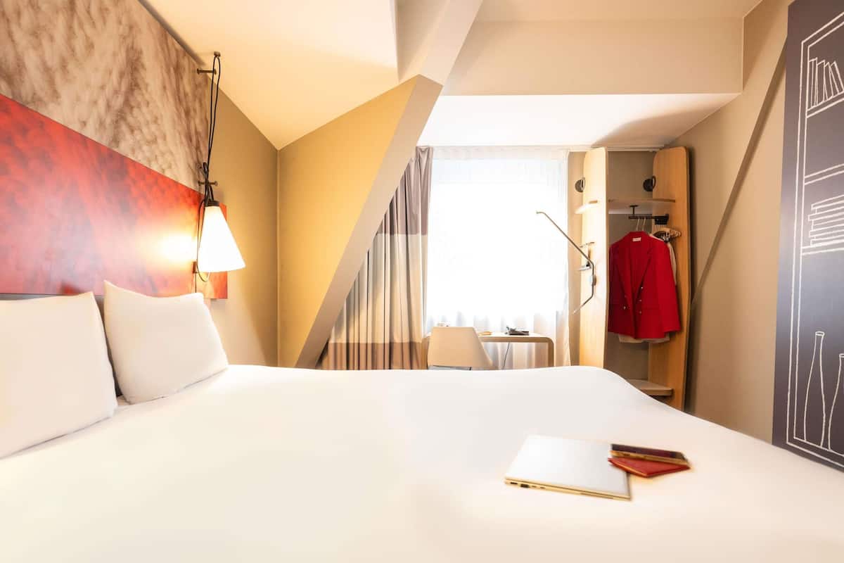 Ein Hotelzimmer mit einem großen Bett, einem Schreibtisch mit Stuhl, einem roten Mantel und Blick ins Freie.