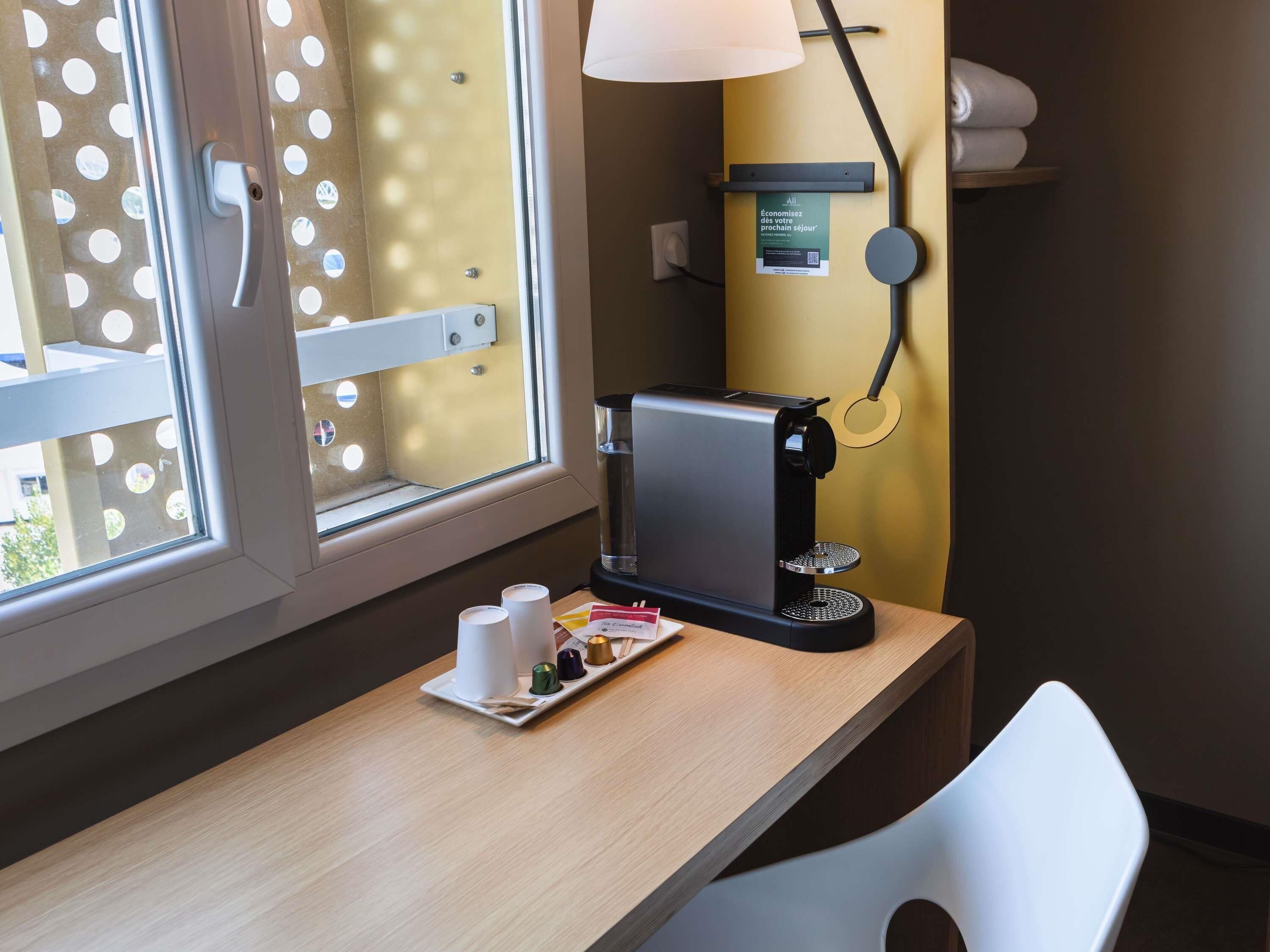 A hotel room with a coffee machine, a small table with cups and a tray, a chair, and a window with a view.