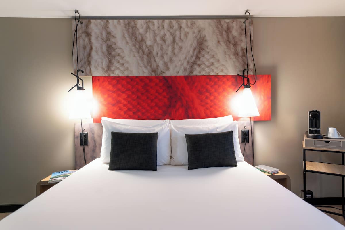 Ein ordentlich bezogenes Bett mit weißen und schwarzen Kissen, einem roten und beigen Kopfteil sowie einem Nachttisch mit Lampe und Kaffeemaschine.