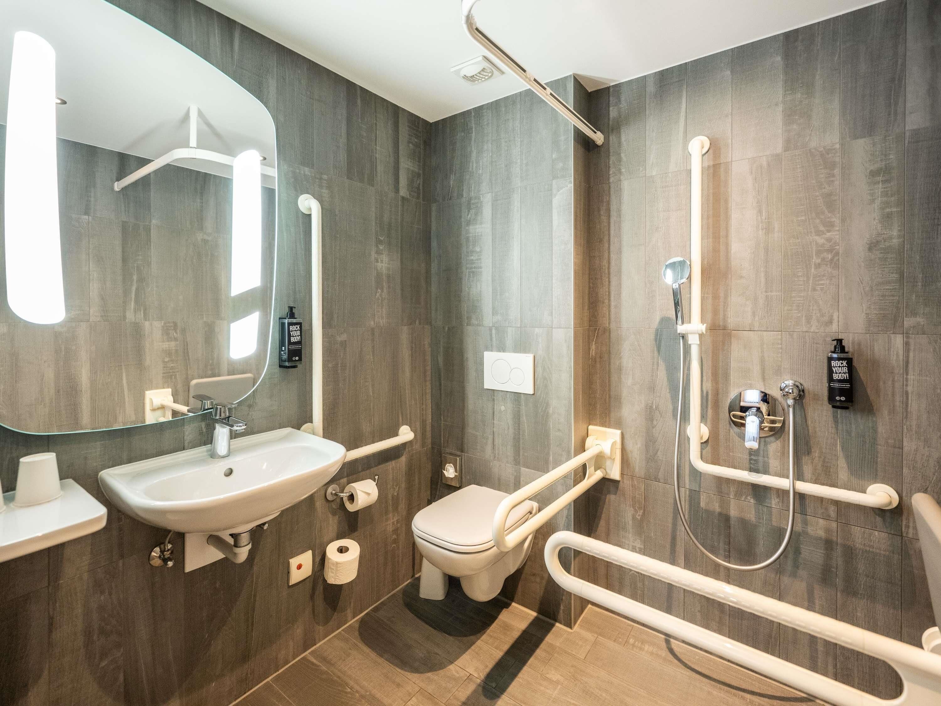 Ein modernes Badezimmer mit Waschbecken, Toilette und Dusche. Es sind Haltegriffe und ein Spiegel vorhanden.