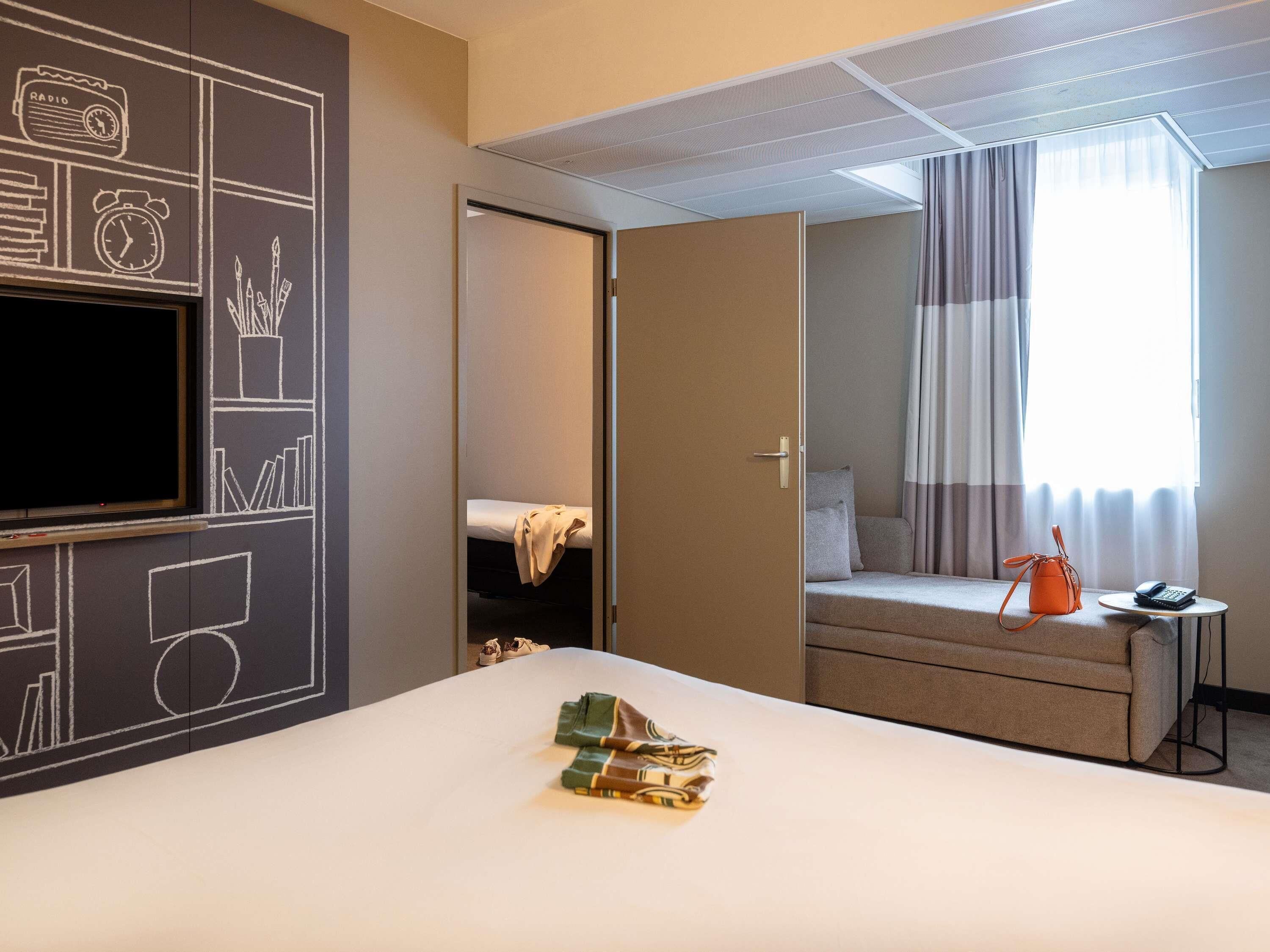 Hotelkamer met een bed, een televisie, een raam met gordijnen, een tafeltje met telefoon en een deur naar een andere kamer.
