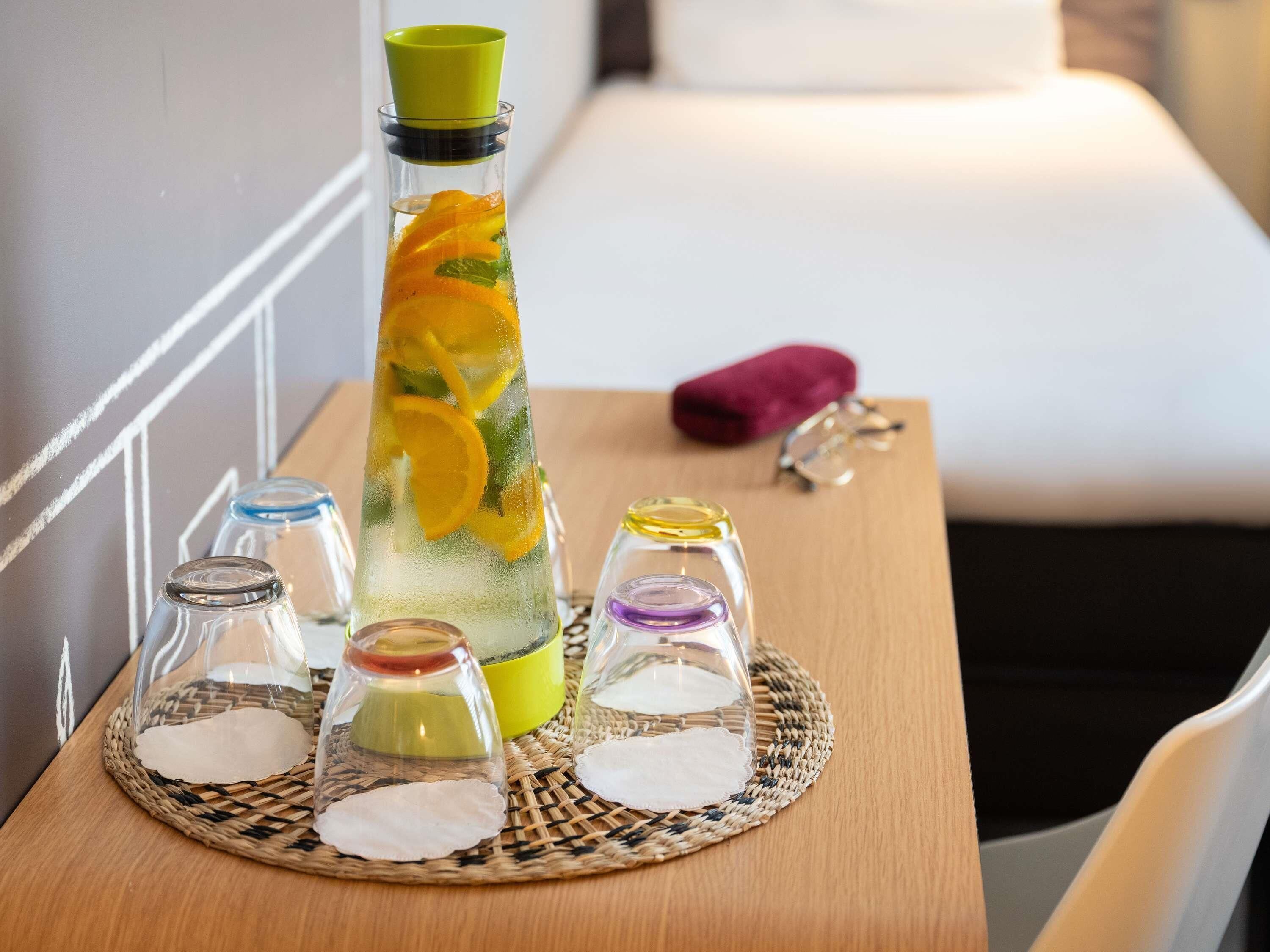 A hotel room with a bed, a bedside table with a water bottle, glasses, and a small vase with orange slices.