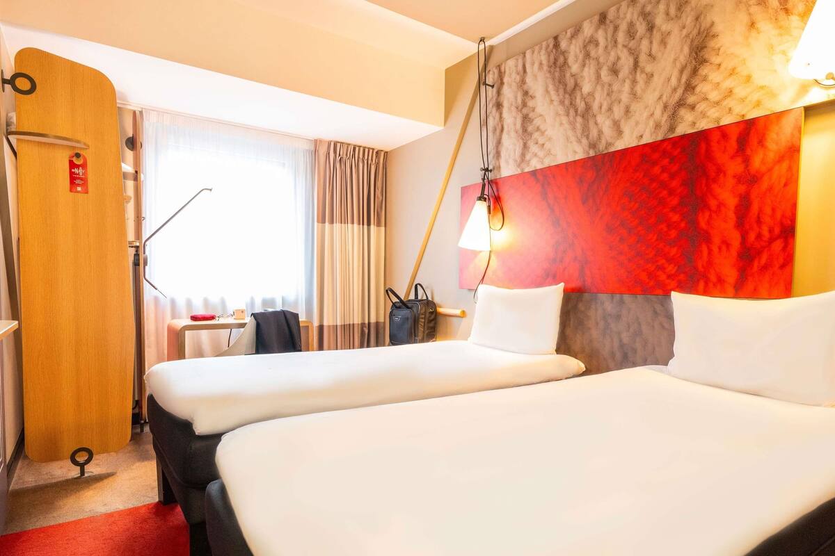 Ein Hotelzimmer mit zwei Betten, einem Holzschrank, einem Schreibtisch mit Stuhl, einem Fenster mit Vorhängen und einer Wand mit einem strukturierten roten und grauen Muster.