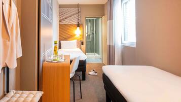 Ein Hotelzimmer mit einem Bett, einem Schreibtisch mit Stuhl, einer kleinen Sitzecke, einer Dusche und einem Fenster mit Vorhängen.