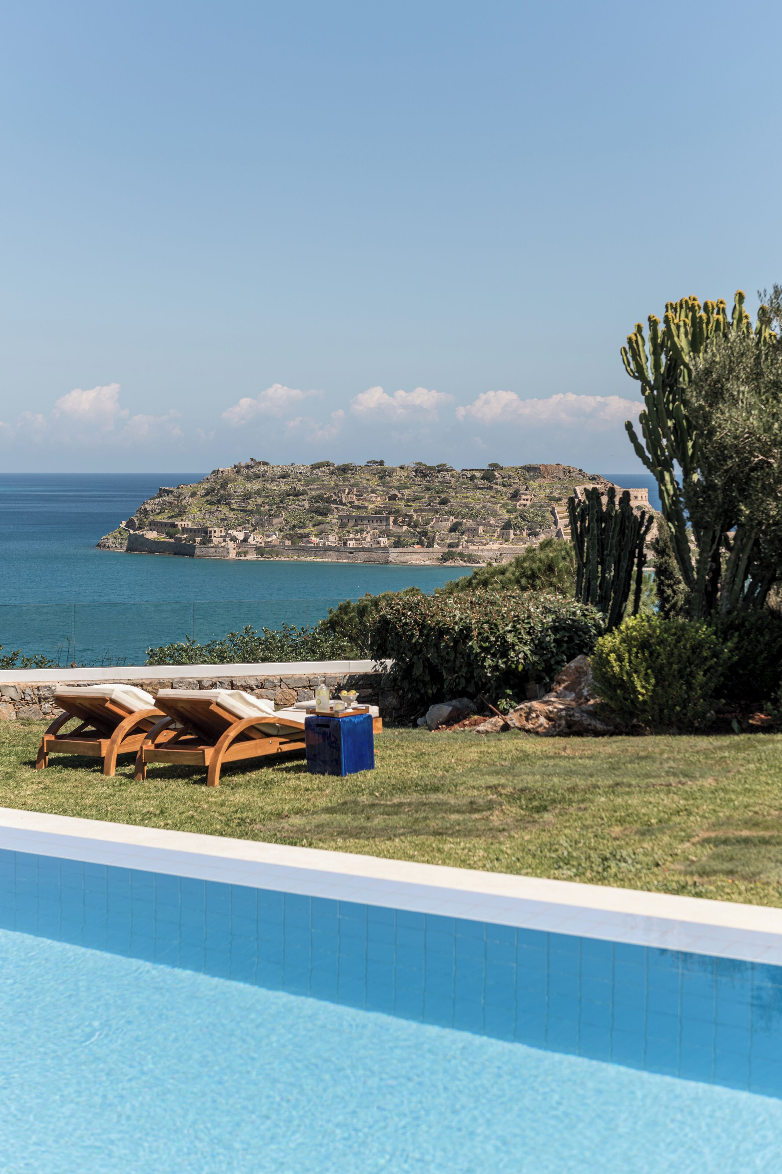 A pool with lounge chairs and a view of the sea and a hill with buildings.