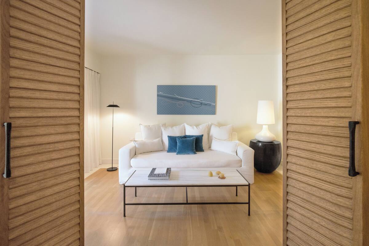 A modern living room with a white sofa, blue pillows, a coffee table, a floor lamp, and a painting on the wall.