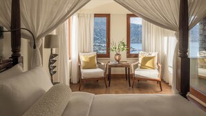 Een slaapkamer met een hemelbed, twee fauteuils, een tafeltje met een vaas, en een uitzicht op een berglandschap door grote ramen.