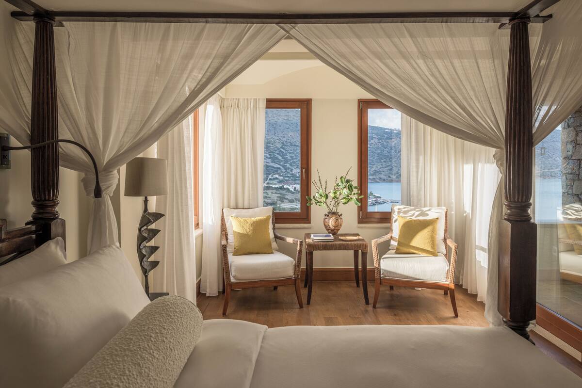 A bedroom with a canopy bed, two armchairs, a small table with a vase, and a view of a mountainous landscape through large windows.
